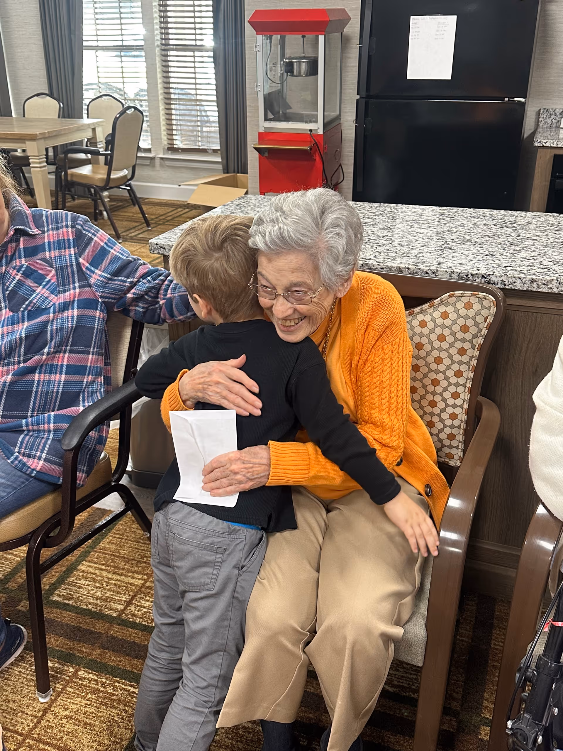 An elderly woman with gray hair and glasses, wearing an orange sweater and beige pants, is sitting on a chair and hugging a young boy dressed in a black shirt and gray pants. They are in a room with a granite countertop, a red popcorn machine, a black refrigerator, and a dining area with tables and chairs in the background.