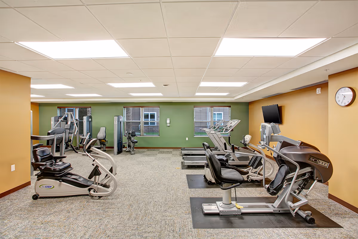 A fitness room with various exercise equipment including recumbent bikes, treadmills, and strength training machines. The room has a green accent wall with three windows, beige walls, and a wall clock. The floor is carpeted, and there is a wall-mounted TV.