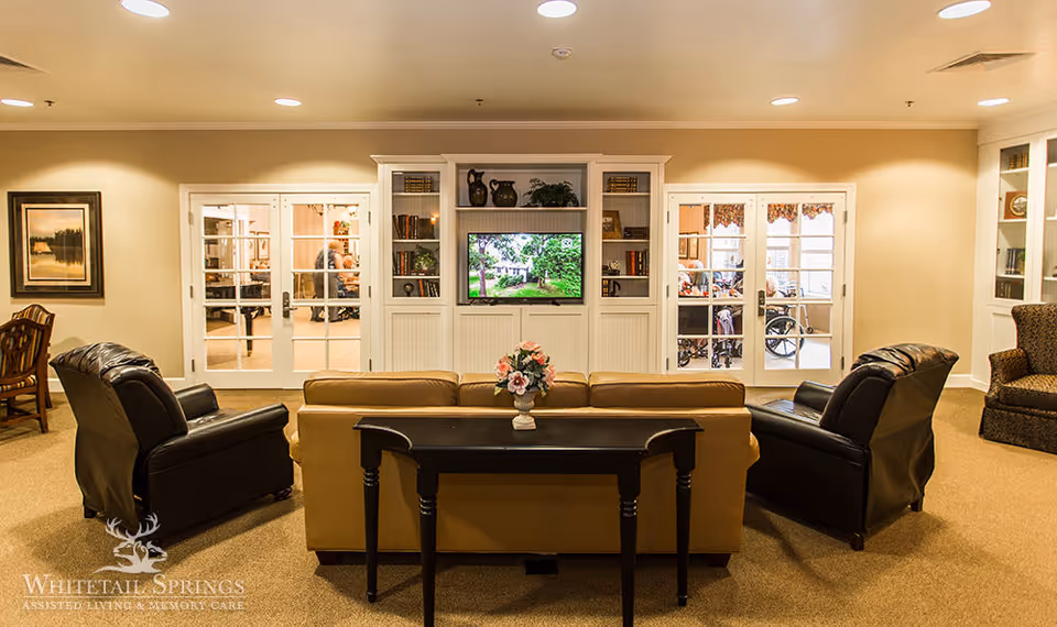 A cozy living room area in a senior living facility featuring a tan sofa with a small black table behind it, two black leather armchairs, a built-in white entertainment center with shelves holding books and decorative items, and a flat-screen TV displaying a nature scene. The room has beige walls, carpeted floors, and French doors leading to another room where people and wheelchairs are visible.