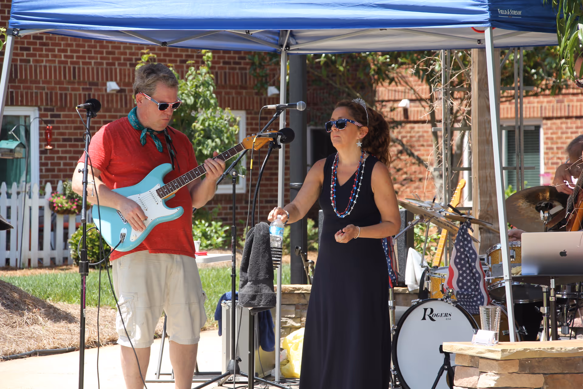 A man playing an electric guitar and a woman standing next to him holding a water bottle under a blue canopy outdoors. The man is wearing a red shirt, beige shorts, sunglasses, and a green scarf around his neck. The woman is wearing a black sleeveless dress, sunglasses, and patriotic bead necklaces. There is a drum set and other musical equipment behind them, with a brick building and greenery in the background.