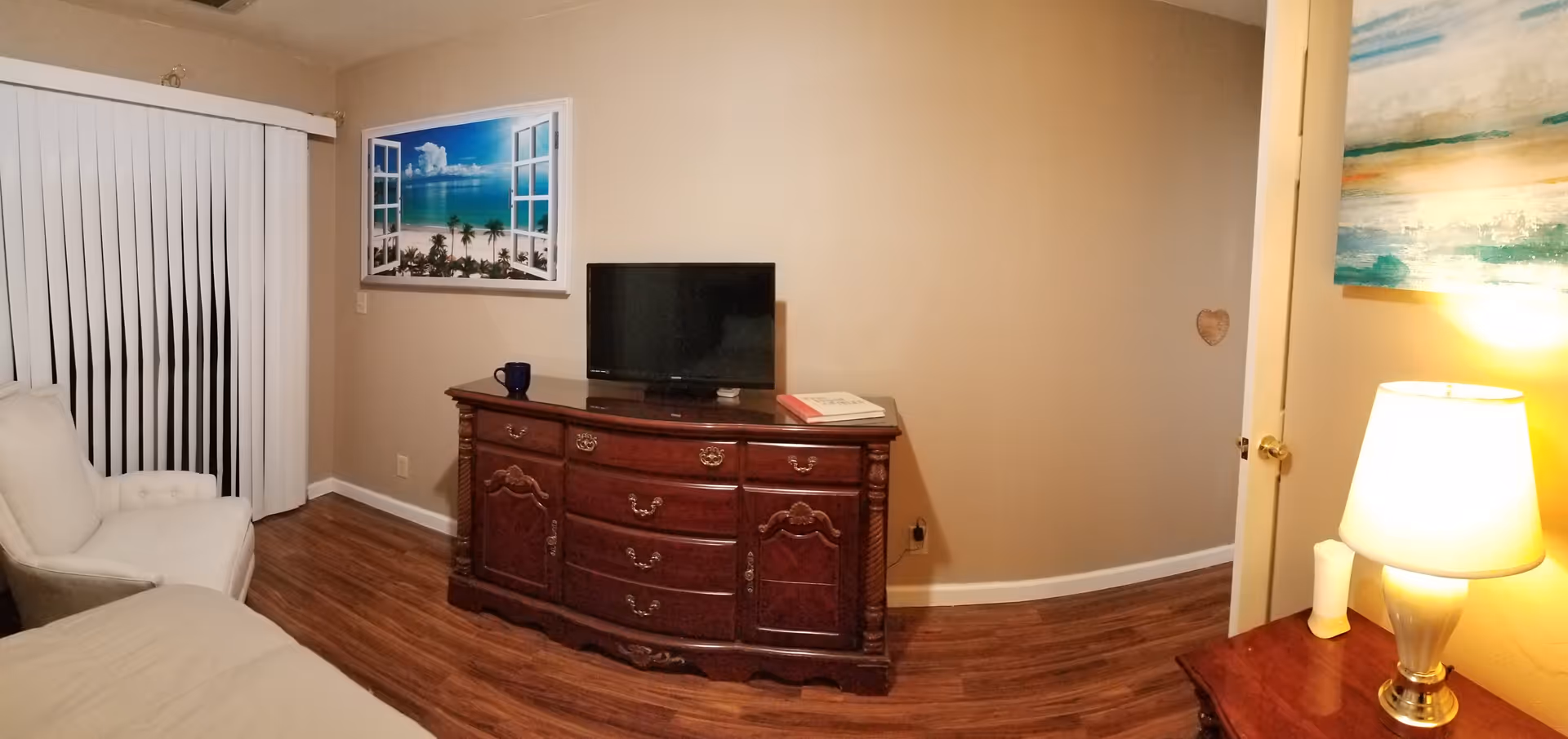 Bedroom with a wooden dresser topped by a TV, an armchair by sliding vertical blinds, and a lit lamp on a nightstand.