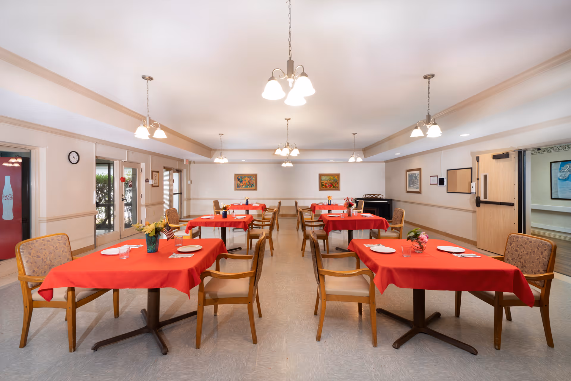 A dining room with several tables covered in red tablecloths, each set with plates, glasses, and small flower arrangements. Wooden chairs with cushioned seats surround the tables. The room is well-lit with multiple ceiling light fixtures and has paintings on the walls. There are double glass doors on the left side and a wooden door on the right.