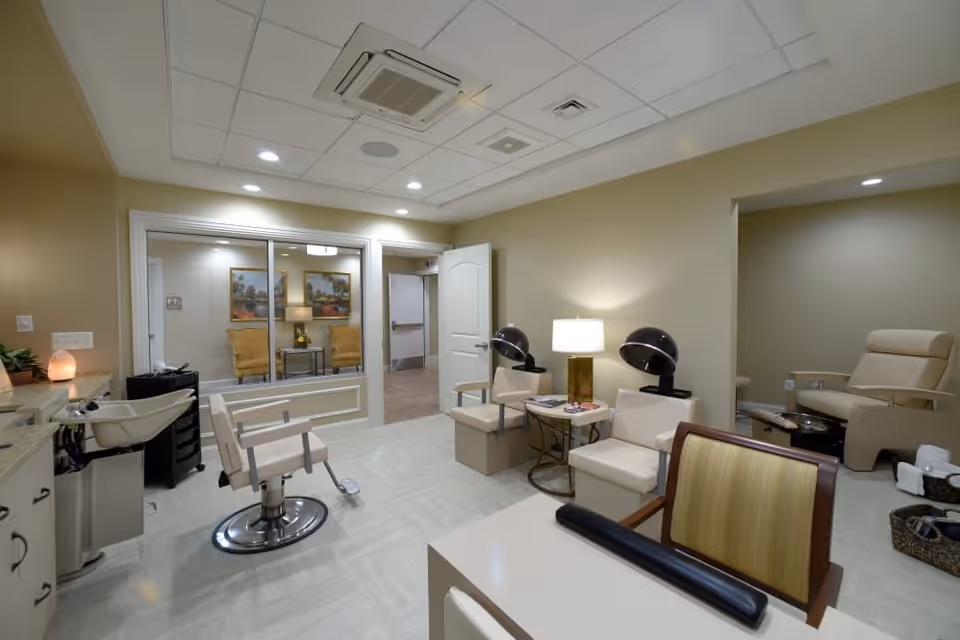 Interior view of a salon area in a senior living facility featuring a styling chair in front of a sink, two hair drying chairs with hooded dryers, a small table with a lamp and magazines, and a pedicure station with a reclining chair. The room has beige walls, a tiled floor, and recessed ceiling lights. A window looks into an adjacent room with two yellow armchairs and framed artwork on the wall.
