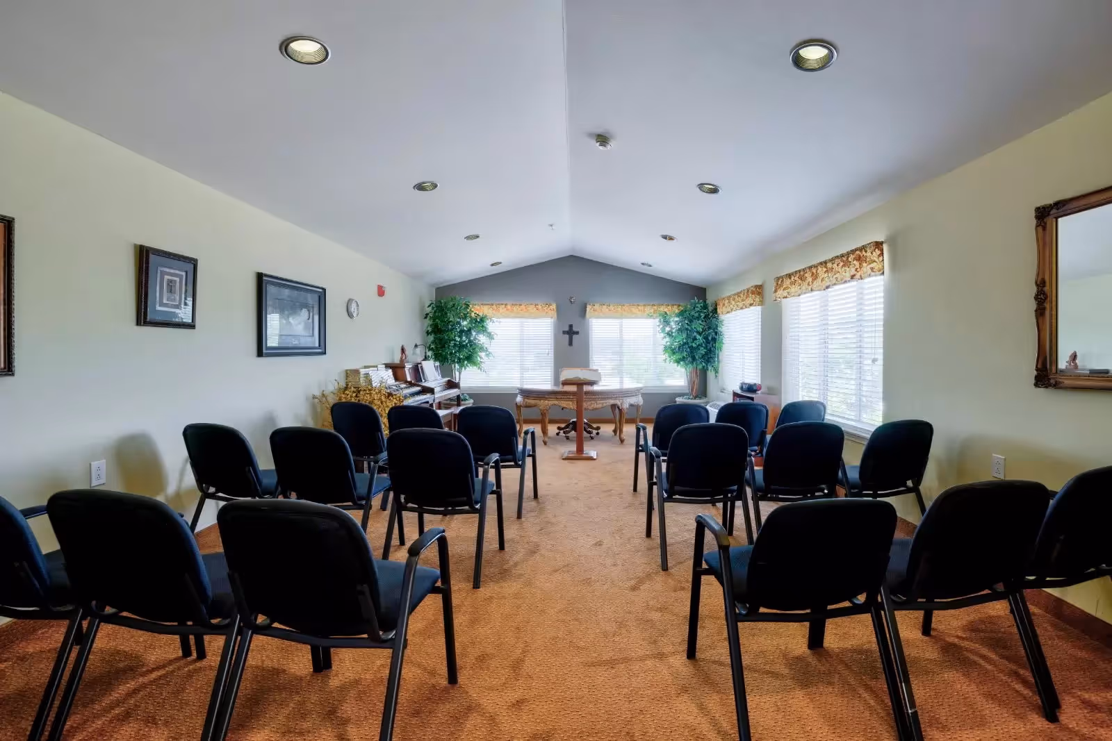 A small chapel or meeting room with rows of black chairs facing a wooden podium and table at the front. The room has beige walls, carpeted floor, framed pictures on the walls, two large windows with floral valances, and two potted plants near the front.