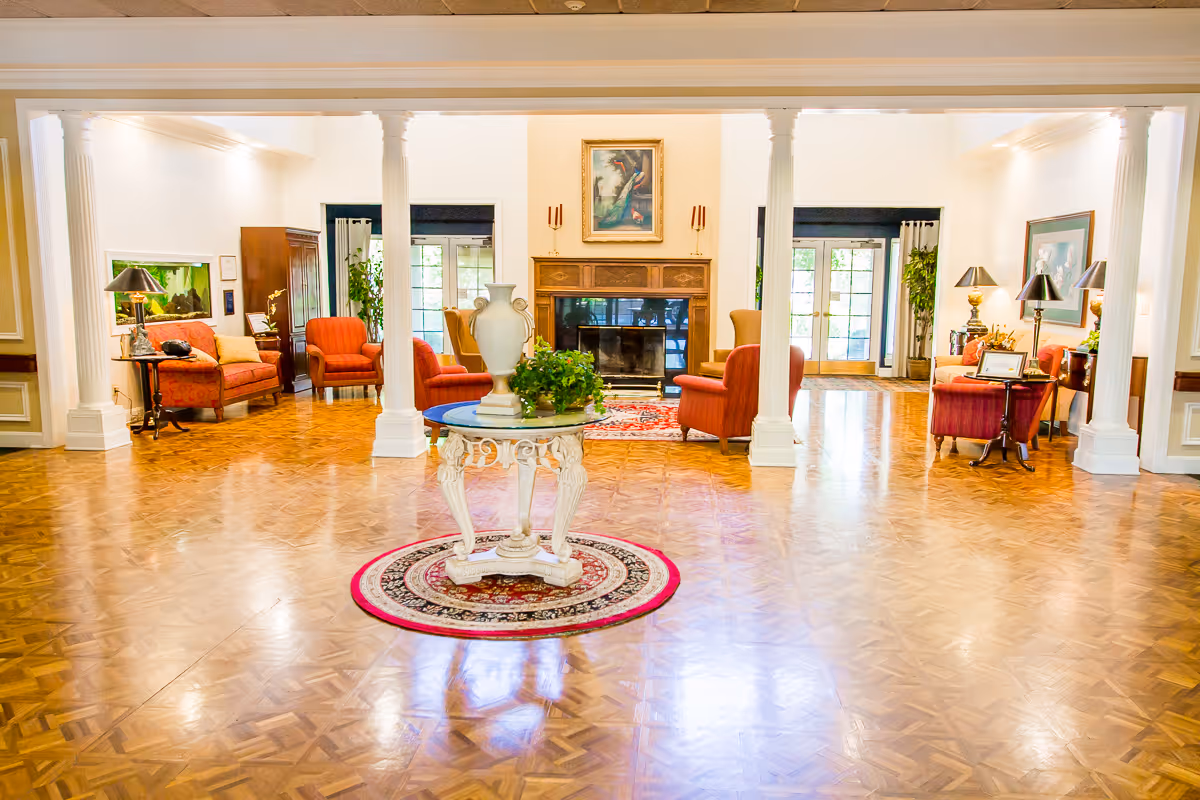 A spacious and elegant living room area with polished wooden parquet flooring, white columns, and a central round table with a decorative vase and greenery. The room features several red upholstered armchairs and sofas arranged around a fireplace with a painting above it. Large windows and glass doors allow natural light to fill the space, and there are various lamps and framed artwork on the walls.