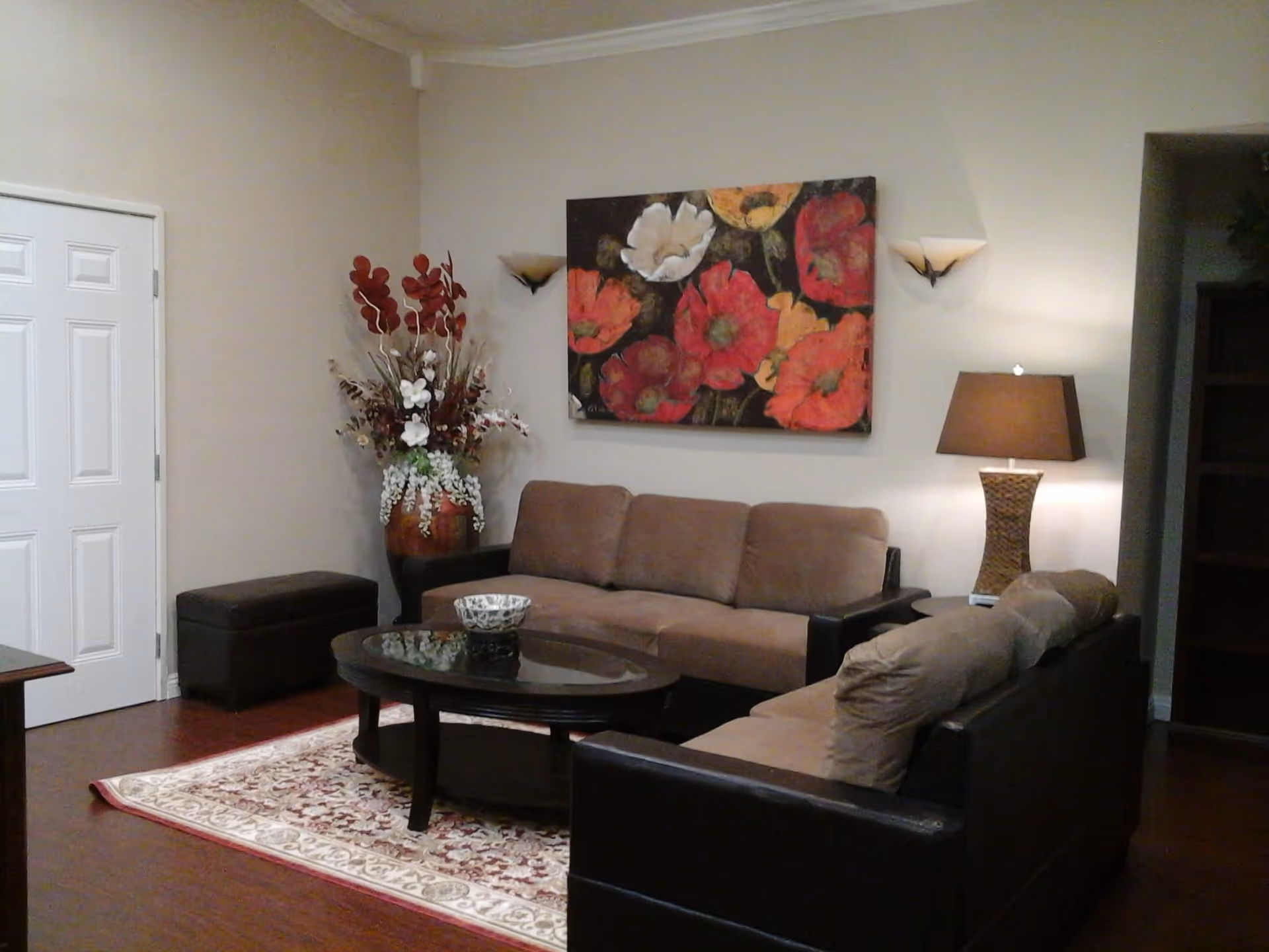 A cozy living room with two brown sofas arranged around a dark wooden oval coffee table on a patterned area rug. There is a large floral painting with red, orange, and white flowers on the wall above one sofa. A tall vase with decorative red and white flowers is placed in the corner near a white door. A table lamp with a brown shade is on a side table next to one sofa, and two wall sconces provide additional lighting.
