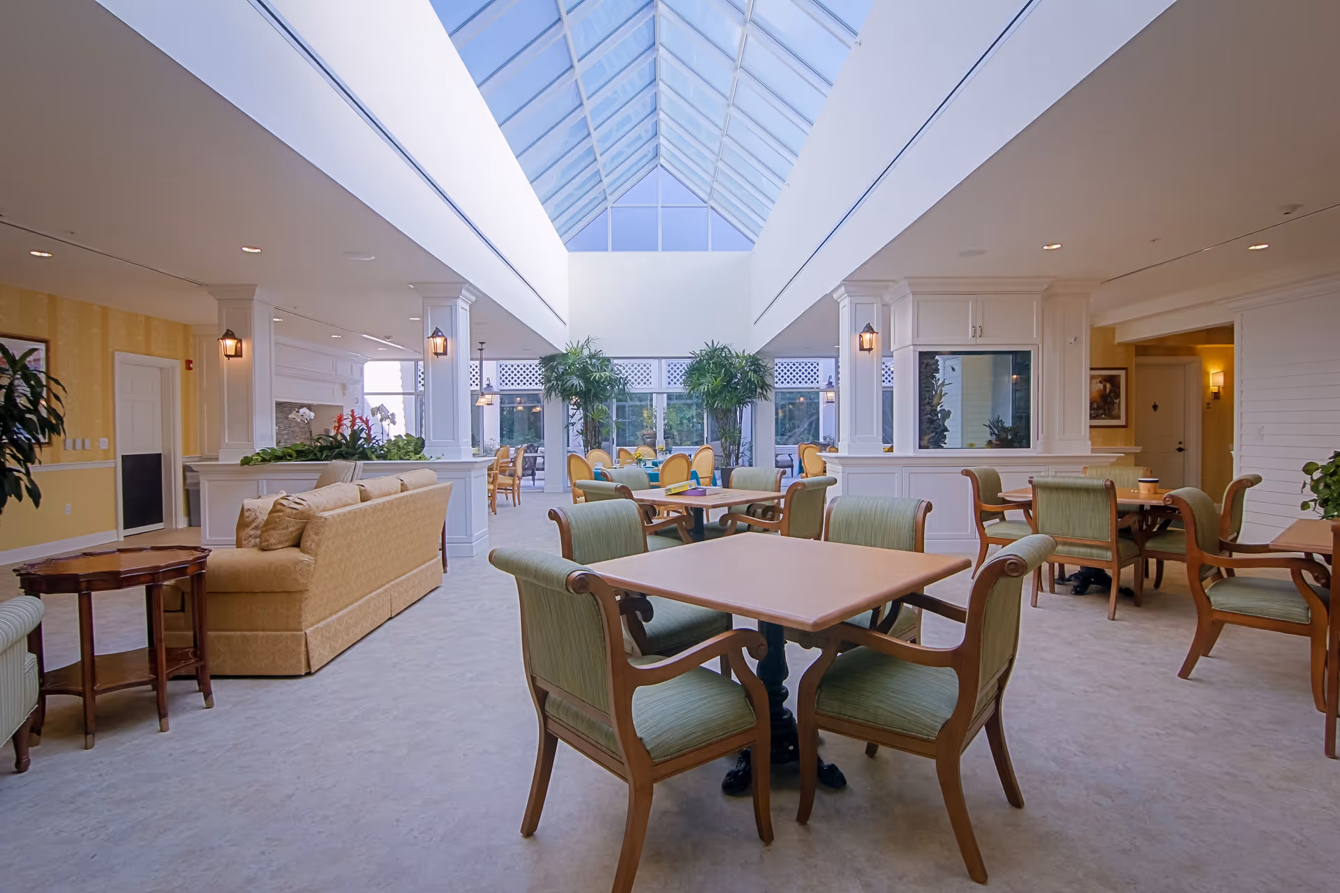 A bright and spacious common area in a senior living facility featuring multiple wooden tables with green cushioned chairs, a beige sofa, potted plants, and a large skylight ceiling allowing natural light to fill the room.
