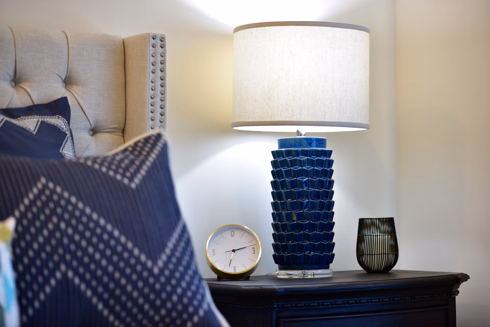 Nightstand with a blue ceramic table lamp, small clock and decorative vase beside a bed with patterned pillows and a tufted headboard.
