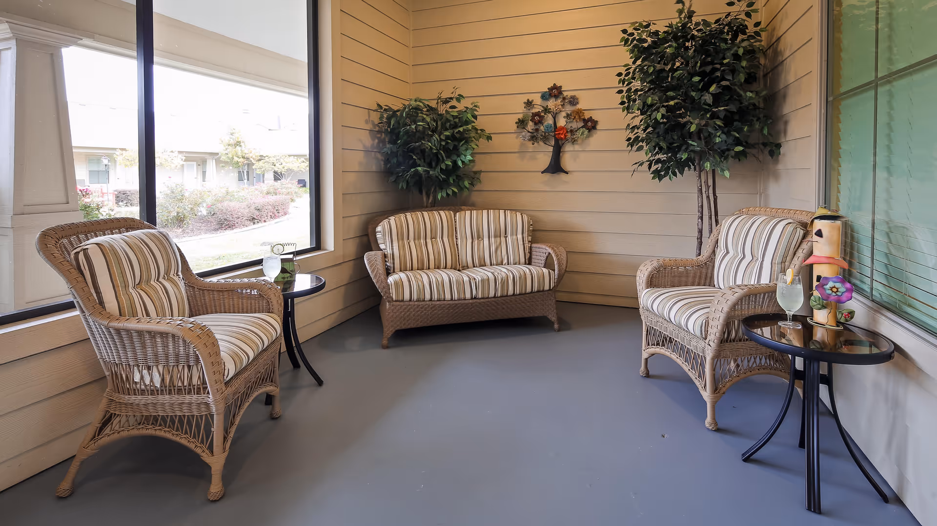 A cozy indoor seating area with three wicker chairs and a wicker loveseat, all with striped cushions. There are two small round glass-top tables, each holding a decorative item and a glass of water with a lemon slice. The walls are light wood paneling, decorated with a metal tree wall art and two large potted plants. Large windows provide a view of the outdoor garden area.