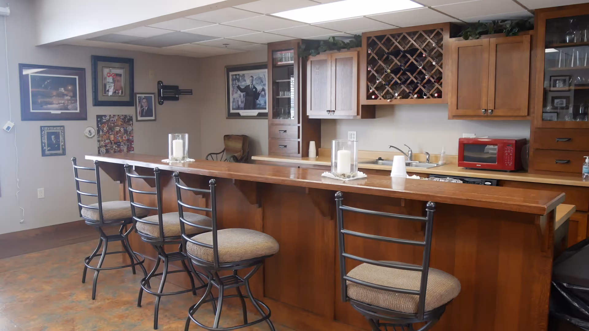 Interior view of a kitchen area with a wooden bar counter and four high chairs with cushioned seats. Behind the counter, there are wooden cabinets, a wine rack with bottles, a red microwave, a sink, and some decorative items. The walls have framed pictures and the floor has a patterned design.