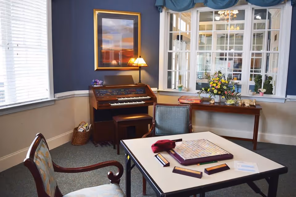A cozy senior living common room with a table and chairs and a board game, an organ against a blue wall, and a console table with flowers beneath interior windows.