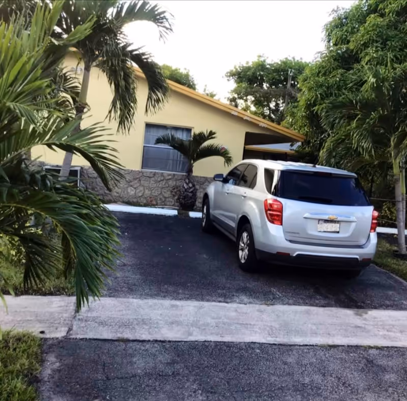 A silver SUV is parked in a driveway in front of a single-story yellow building with a stone lower facade. The area is surrounded by palm trees and other greenery.