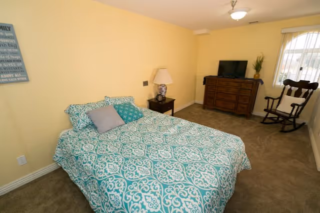 A bedroom with a bed covered in a teal and white patterned comforter and pillows. There is a wooden nightstand with a lamp next to the bed, a wooden dresser with a TV on top, a rocking chair with a cushion, and a window with sheer curtains letting in natural light. The walls are painted light yellow and the floor is carpeted.