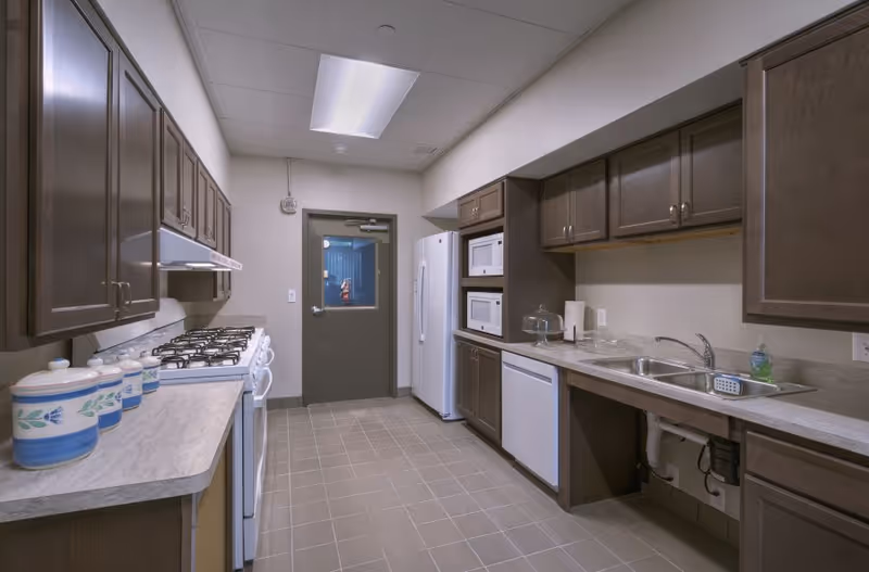 A clean, well-lit kitchen with brown cabinets, a white refrigerator, two microwaves stacked in a cabinet, a dishwasher, a double sink, a gas stove with four burners, and a countertop with decorative canisters. The kitchen has tiled flooring and a door with a window at the end.