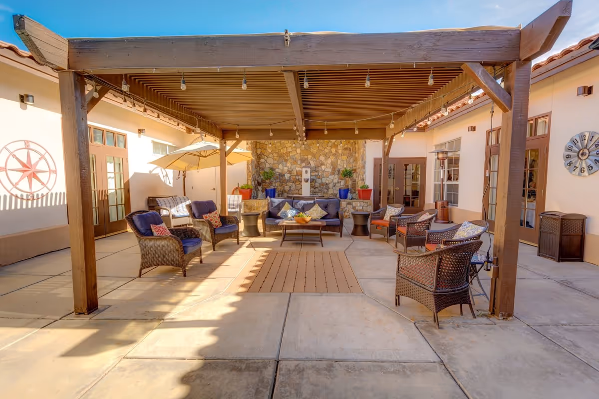 Outdoor patio area at The Auberge at Scottsdale featuring a wooden pergola with string lights, wicker chairs with cushions arranged around a coffee table, a stone wall with potted plants, and umbrellas providing shade.