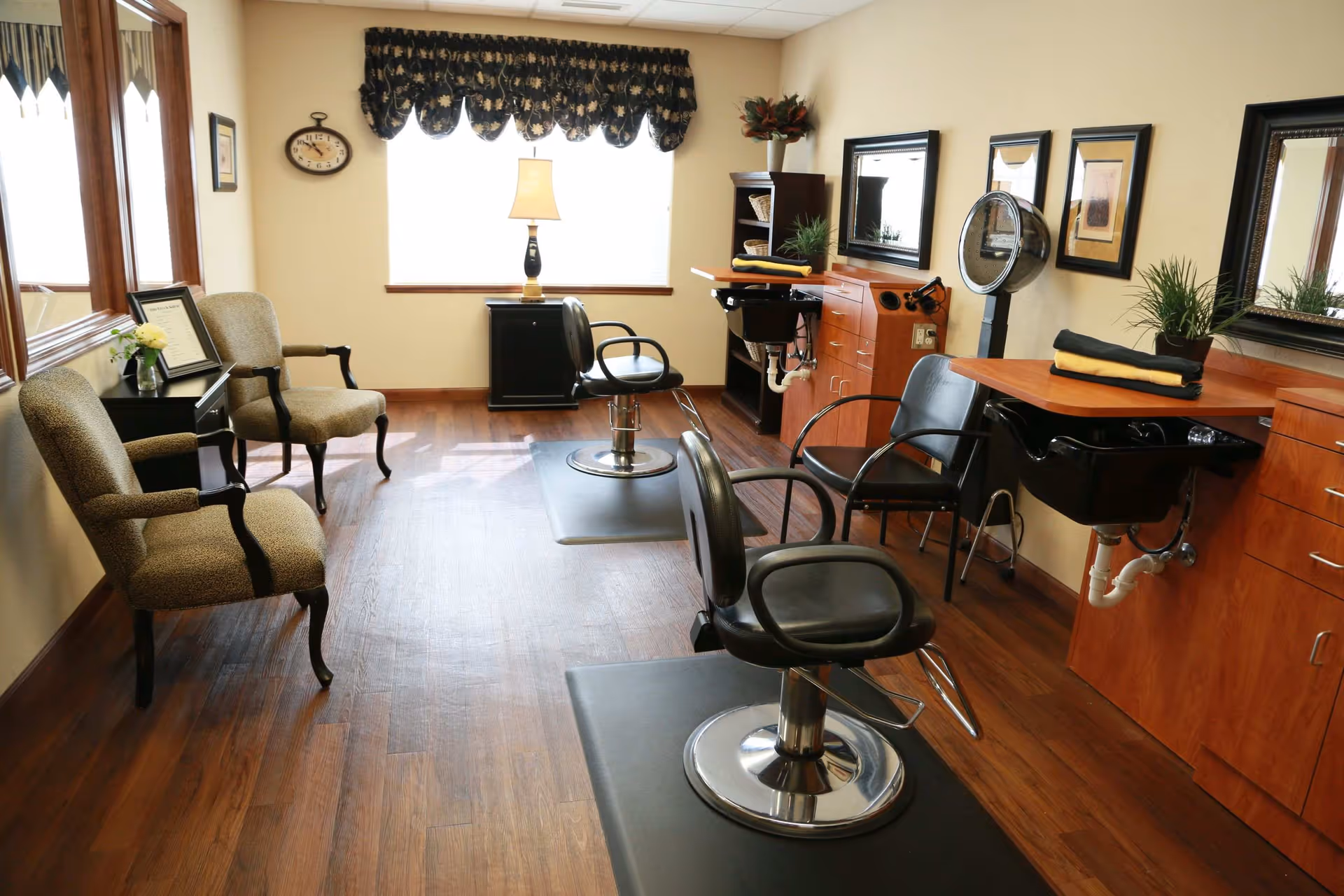 Interior view of a salon area with two black styling chairs in front of wooden counters with sinks and mirrors. There are two upholstered armchairs along the left wall, a small table with a flower vase, a clock on the wall, and a window with a decorative valance letting in natural light.