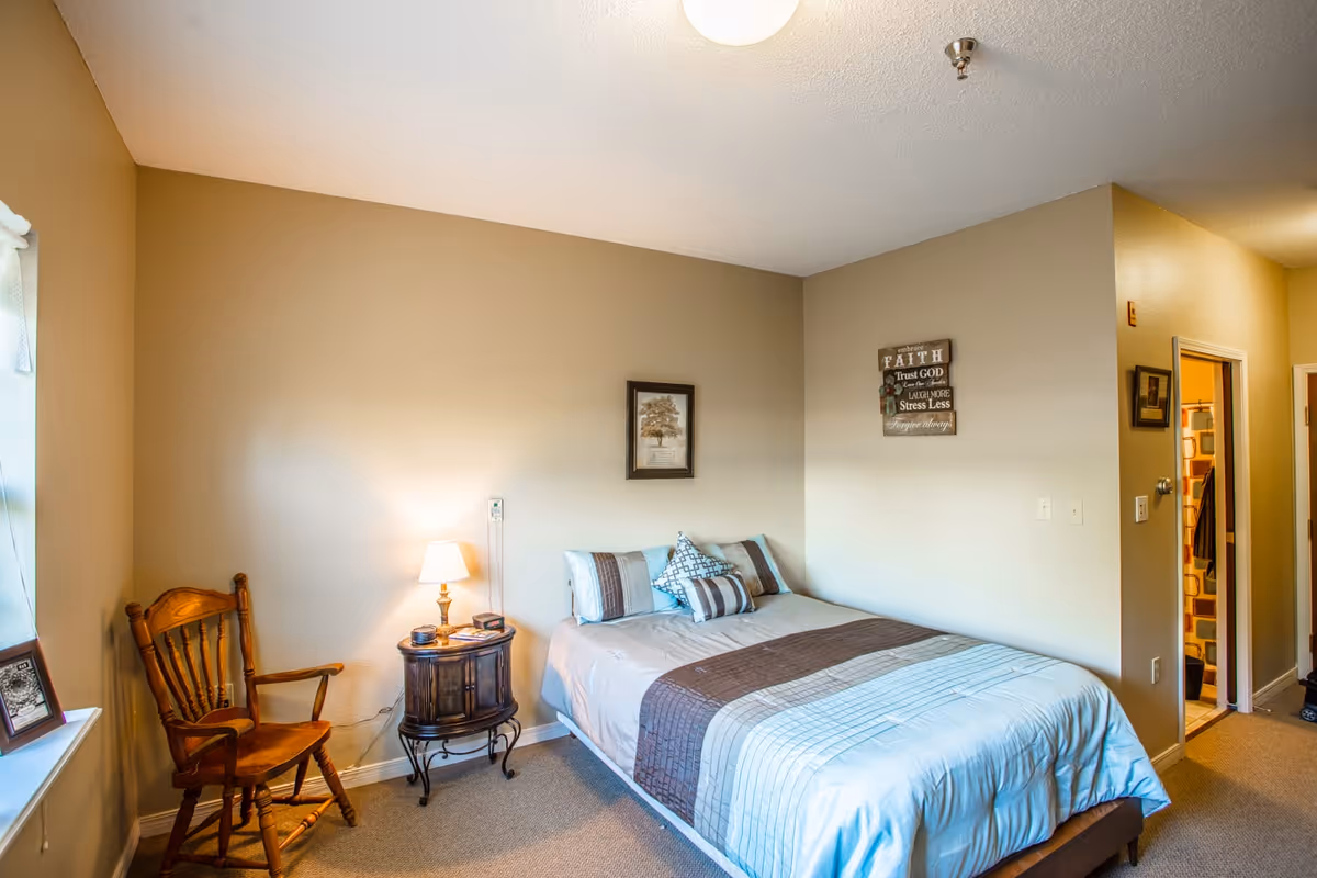 A cozy bedroom with a bed made up with a striped comforter and several pillows. Next to the bed is a small round wooden nightstand with a lamp and some items on it. A wooden chair is placed near the window on the left side. The walls are painted beige and decorated with two framed pictures. A doorway on the right leads to a bathroom with a shower curtain visible.