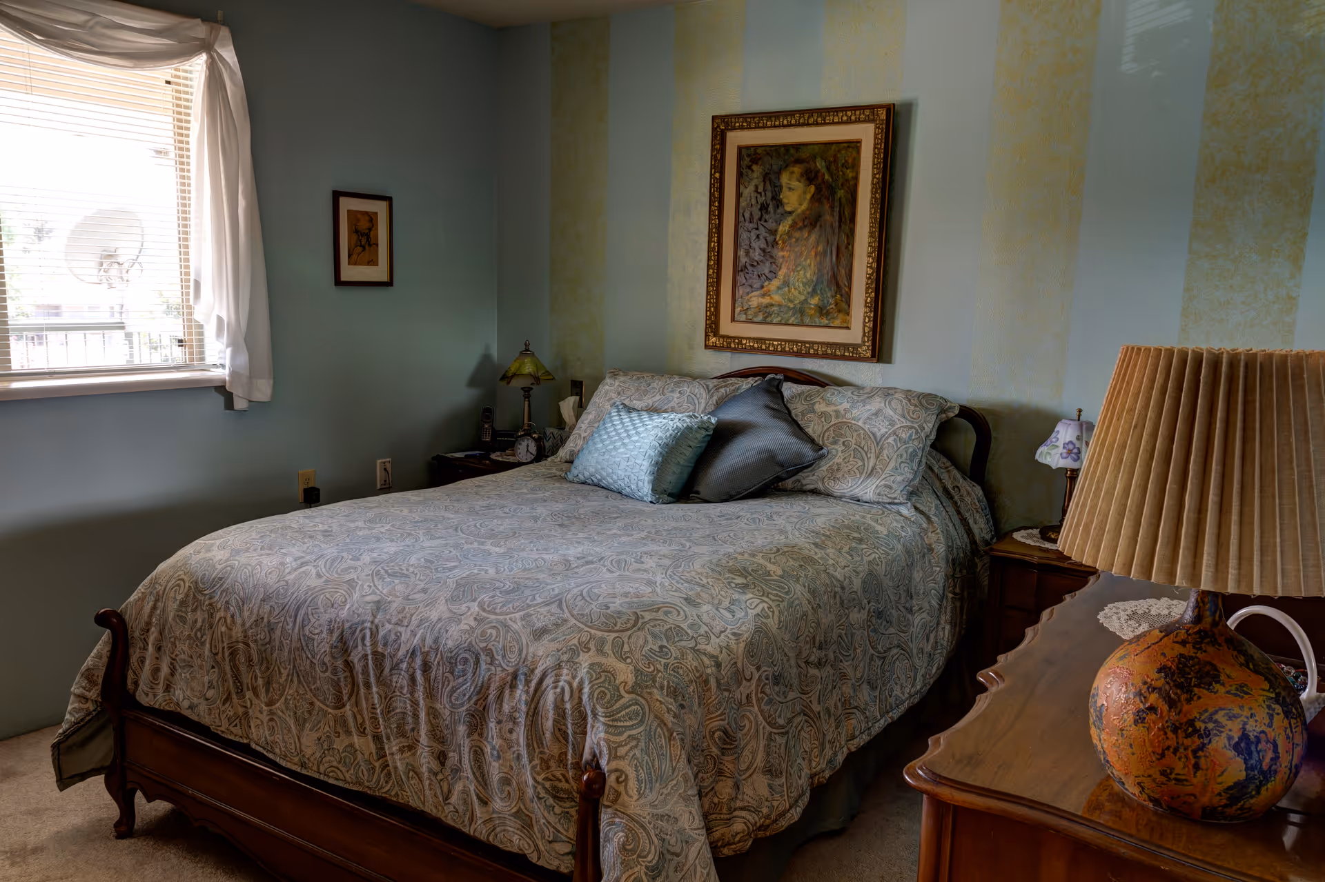 A cozy bedroom with a wooden bed frame and a paisley-patterned bedspread. There are three pillows on the bed, two with a paisley design and one solid blue. The walls are painted with vertical stripes in light blue and yellow. A framed painting of a woman hangs above the bed. On the left side, there is a window with white curtains partially open, letting in natural light. Two nightstands flank the bed, each with a lamp, and a wooden dresser with a large, textured lamp is visible in the foreground.