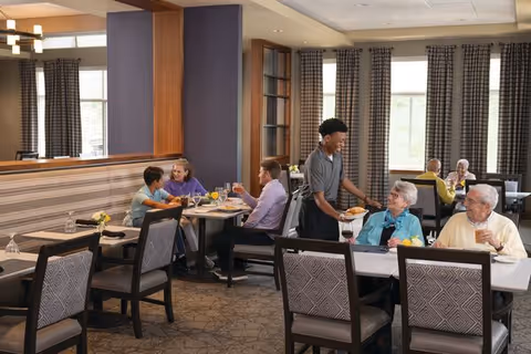Residents eating and conversing in a senior living community dining room while a server brings plates.