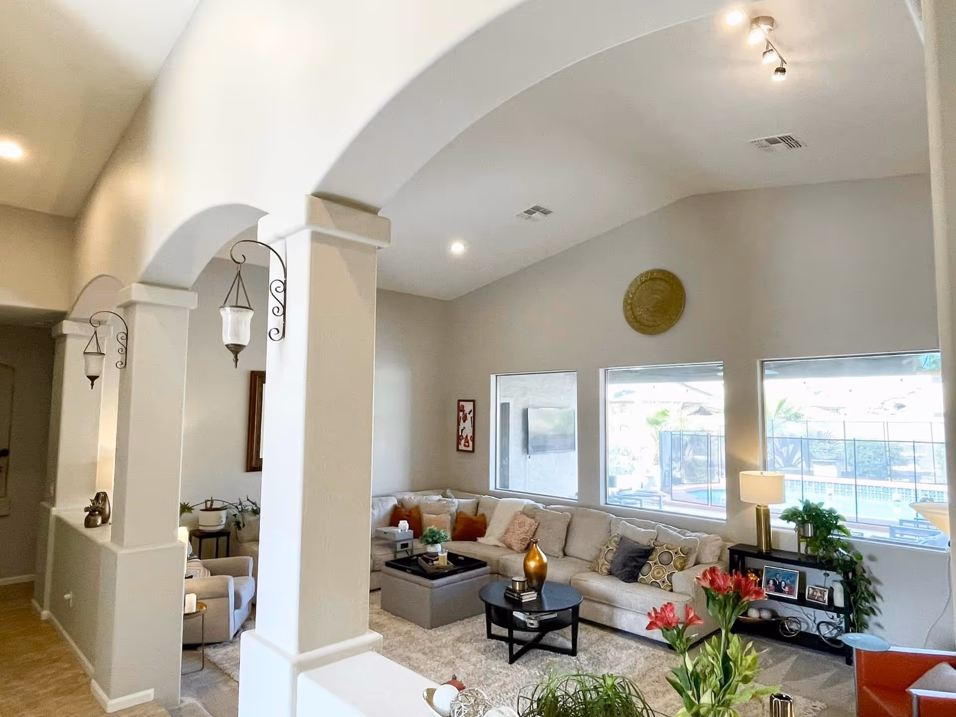 A bright living room with high vaulted ceilings and three large windows showing an outdoor pool area. The room features a large beige sectional sofa with multiple cushions, a round black coffee table with decorative items, a side table with a lamp and plants, and a soft area rug. The room is separated by white pillars with decorative hanging lanterns.