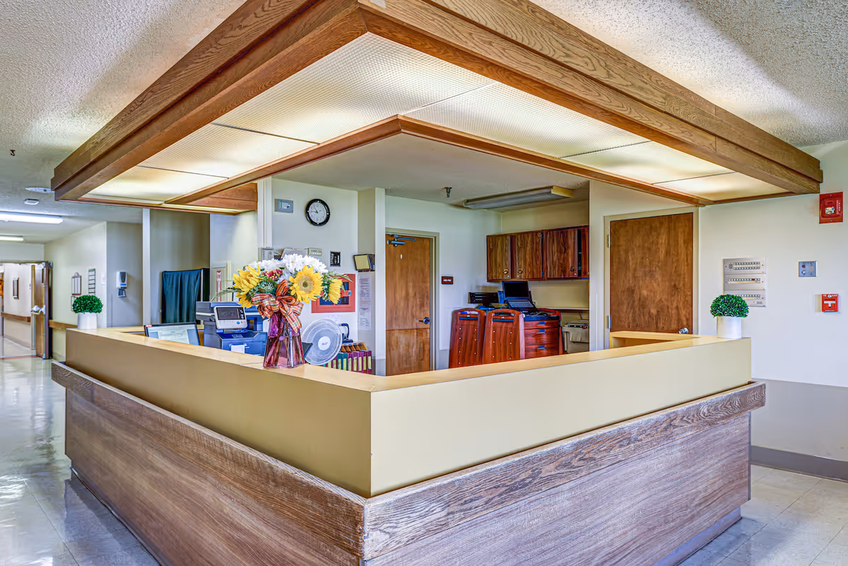 Reception desk area in a senior living facility with a bouquet of flowers in a vase on the counter, office equipment, wooden cabinets, and doors in the background. The ceiling has a wooden frame with fluorescent lighting, and there are small potted plants on the counter edges.