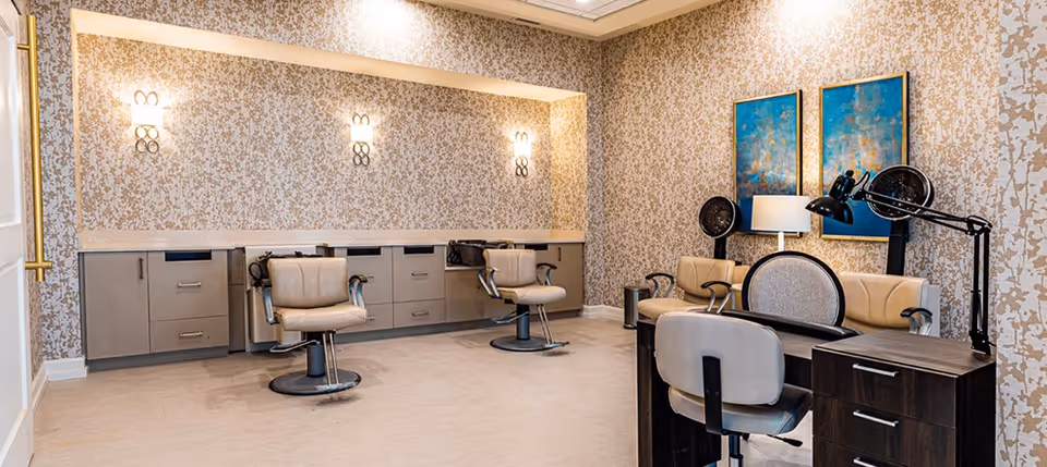 Interior view of a salon area in a senior living facility with beige salon chairs, hair dryers, a desk with a lamp, and two blue and gold abstract paintings on the wall. The walls have a patterned wallpaper and the floor is light-colored.