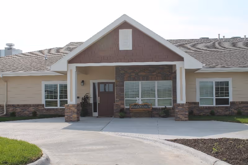 Front exterior view of a single-story building with a covered entrance supported by two columns. The building has beige siding with brick accents and a brown shingled roof. There are windows on either side of the entrance and a small landscaped area with grass and mulch in front.