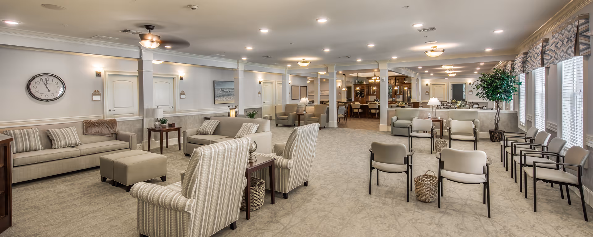 A spacious and well-lit senior living facility common area with multiple seating arrangements including sofas, armchairs, and chairs lined up near large windows with patterned valances. The room features neutral tones, carpeted flooring, ceiling fans, recessed lighting, and decorative elements such as a wall clock, framed artwork, and potted plants. In the background, there is a kitchen area with bar stools and dining tables.