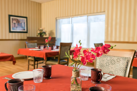 Dining room with tables covered in red tablecloths, set with plates, glasses, and dark-colored mugs. A small vase with bright pink flowers is placed on the nearest table. The room has striped wallpaper, a large window with blinds, and framed artwork on the wall.