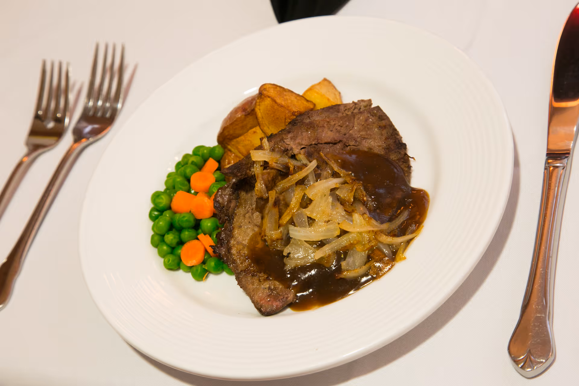 A white plate with a serving of cooked meat topped with sautéed onions and brown gravy, accompanied by cooked green peas and sliced carrots, and roasted potato wedges. The plate is set on a white tablecloth with two forks on the left and a knife on the right.