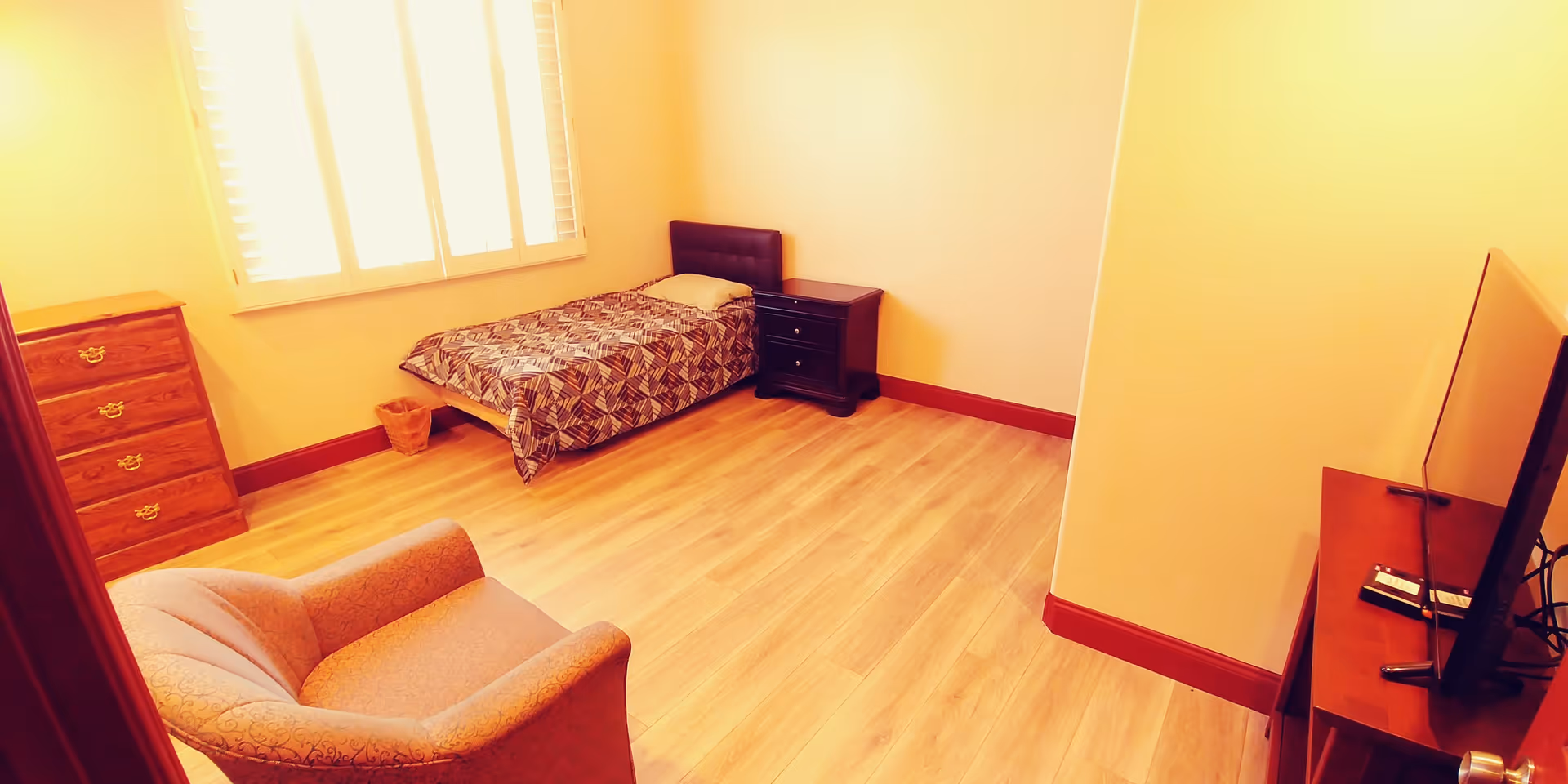 A simple bedroom with a single bed covered in a patterned bedspread, a wooden dresser, a nightstand, an upholstered armchair, and a flat-screen TV on a wooden stand. The room has light-colored walls, wood flooring, and a window with white shutters letting in natural light.