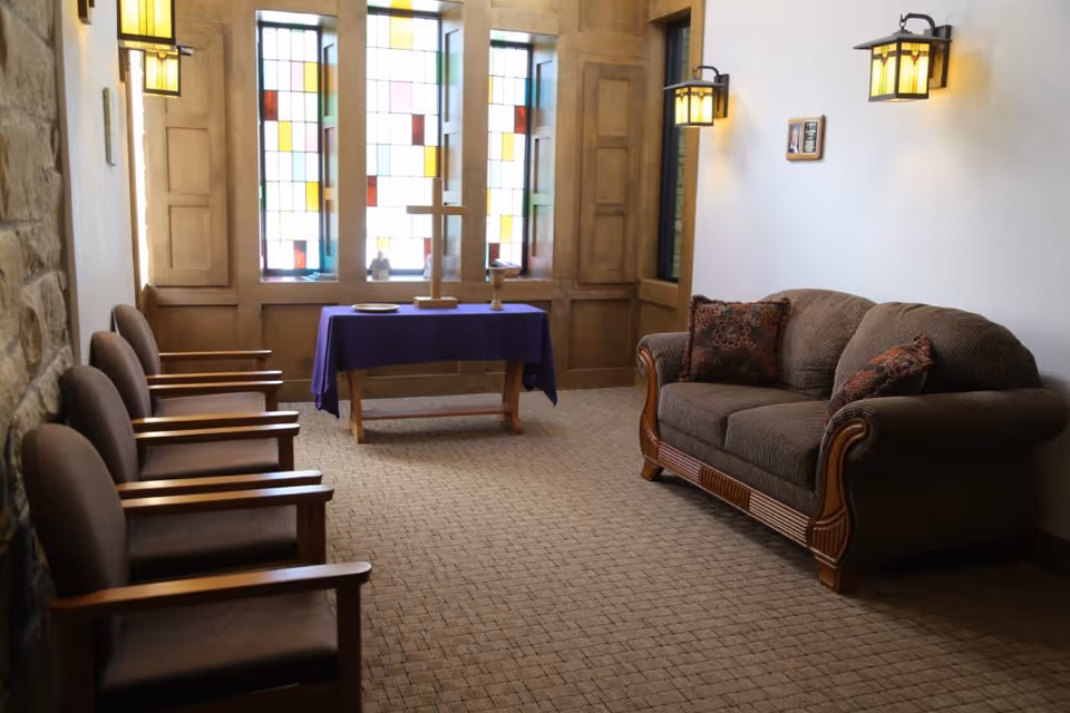 A small chapel or prayer room with a brown upholstered couch on the right and four wooden chairs with brown cushions on the left. At the far end, there is a wooden table covered with a purple cloth, holding a wooden cross and other religious items. Behind the table are stained glass windows with colorful geometric patterns. The room has wall-mounted lantern-style lights and a carpeted floor.