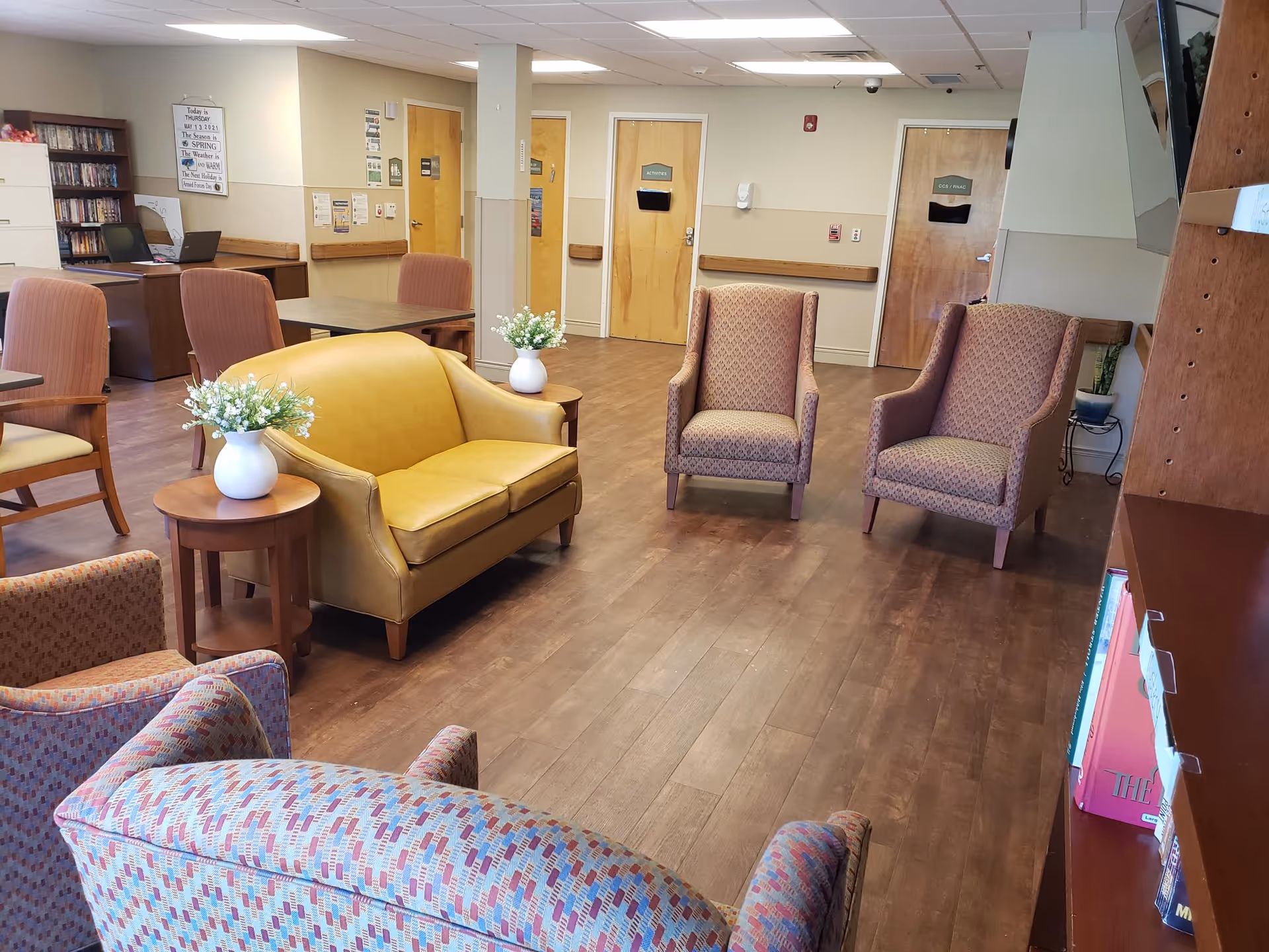 A senior living facility lounge with armchairs, a yellow loveseat, side tables with vases of flowers, bookshelves, and doors along the back wall.