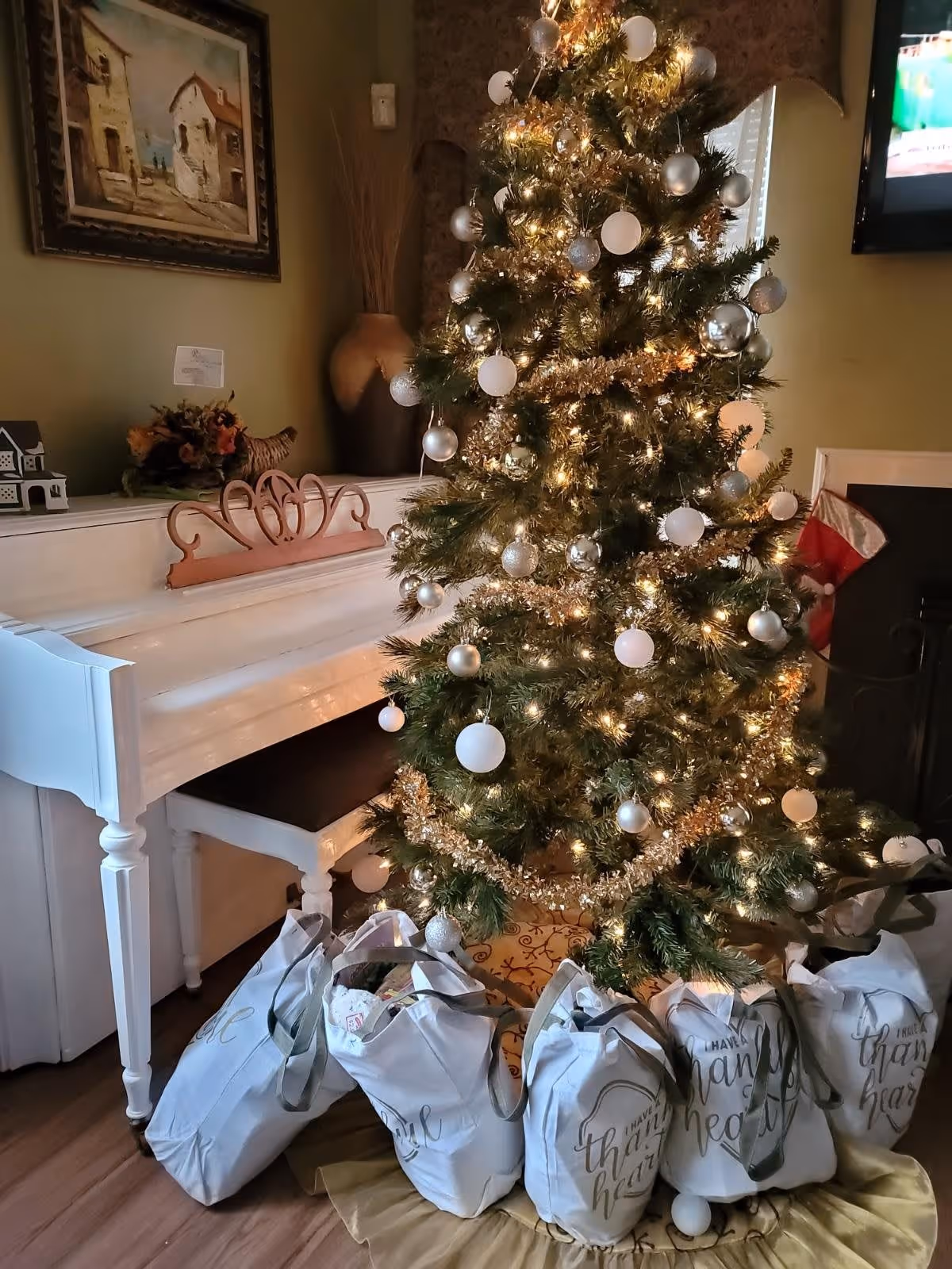 A decorated Christmas tree with white and silver ornaments and lights stands next to a white piano in a cozy room. Several white gift bags with the words 'Have a thankful heart' are placed under the tree. A painting hangs on the wall above the piano, and a television is mounted on the wall to the right.