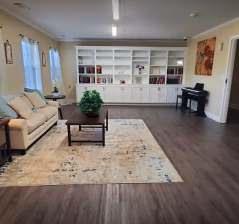 Spacious communal living room with a sofa, coffee table on a rug, built-in bookshelves, and a piano.