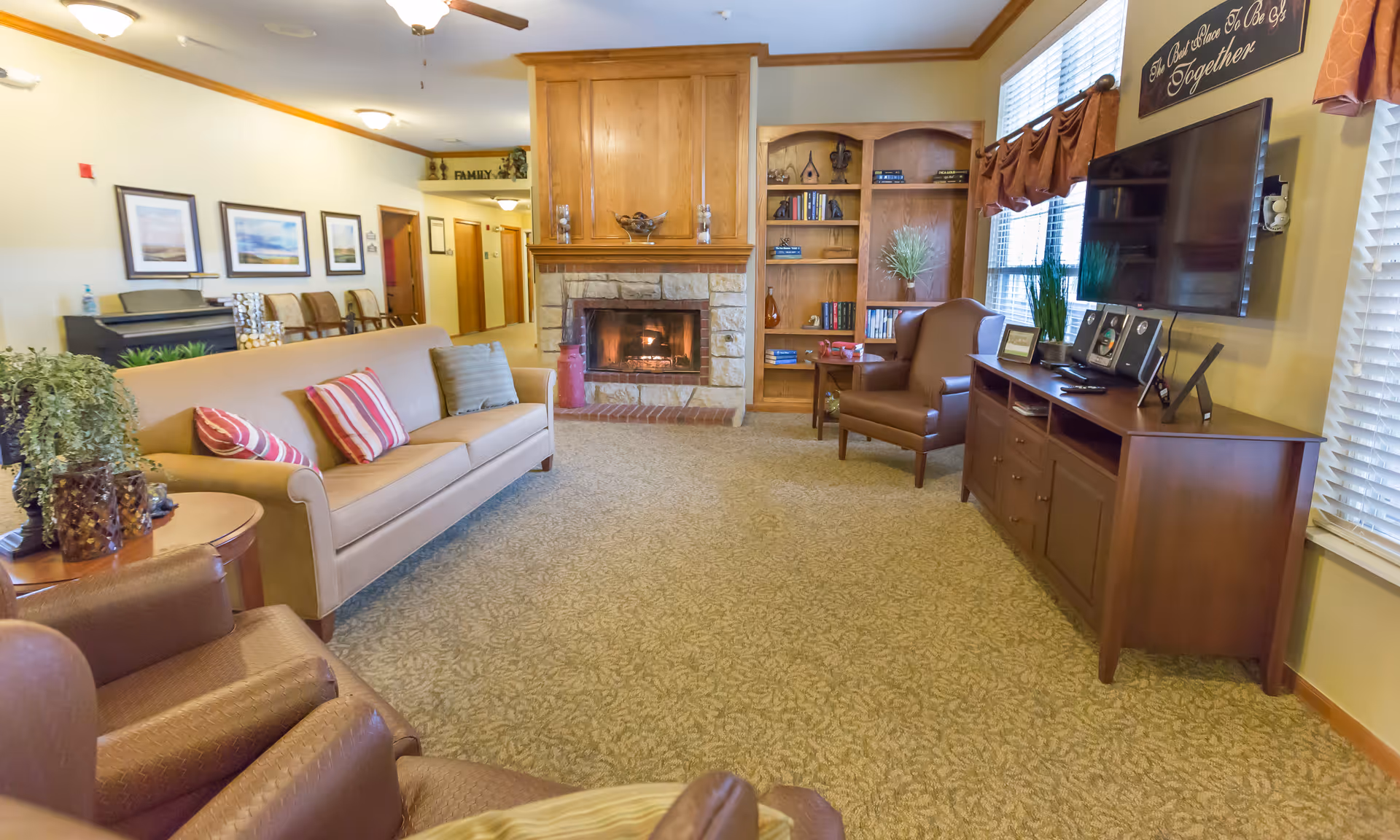 A cozy living room in a senior living facility featuring a beige sofa with striped pillows, two brown armchairs, a stone fireplace with a wooden mantel, built-in wooden bookshelves, a flat-screen TV mounted on the wall, and large windows with blinds and valances. The room is carpeted and decorated with plants, framed pictures, and a sign above the TV that reads 'The Best Place To Be Is Together.'