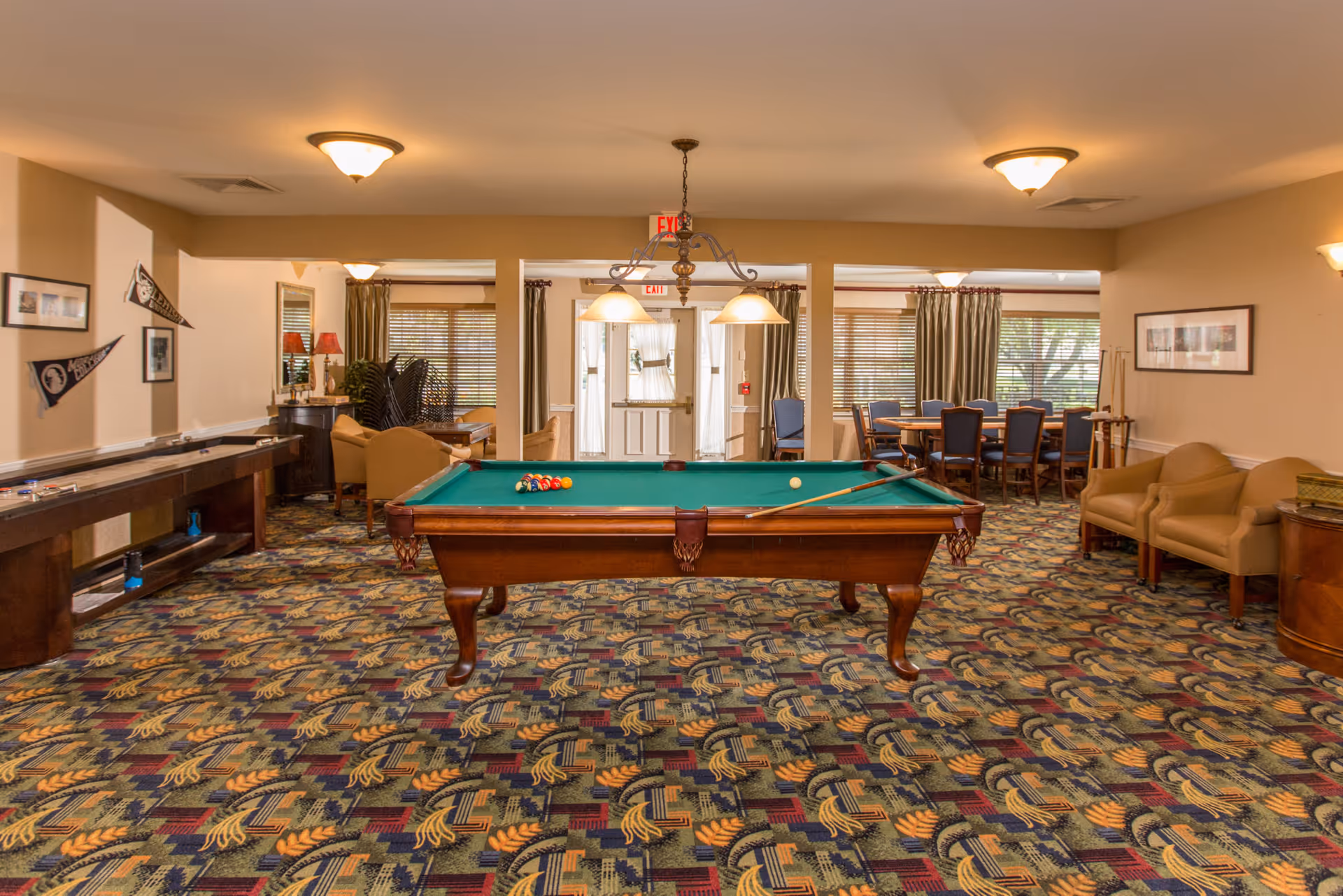 A spacious recreational room with a pool table in the center, a shuffleboard table to the left, and a seating area with chairs and tables in the background. The room has patterned carpet, beige walls, and multiple light fixtures on the ceiling. There are windows with blinds and curtains, and framed pictures and pennants on the walls.