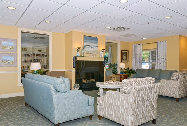 A cozy living room area in a senior living facility with light blue and patterned armchairs and sofas arranged around a white round coffee table. The room features a black fireplace with decorative items on the mantel, soft yellow walls, framed artwork, and large windows with white curtains allowing natural light to enter.