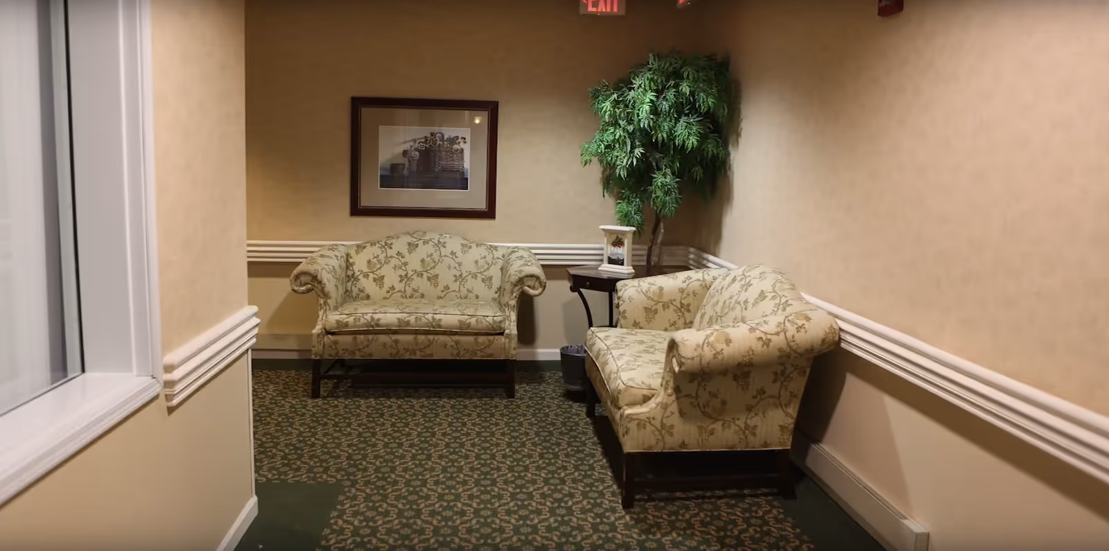 Small seating area in a carpeted hallway with two patterned loveseats, a side table, framed art and a potted plant.