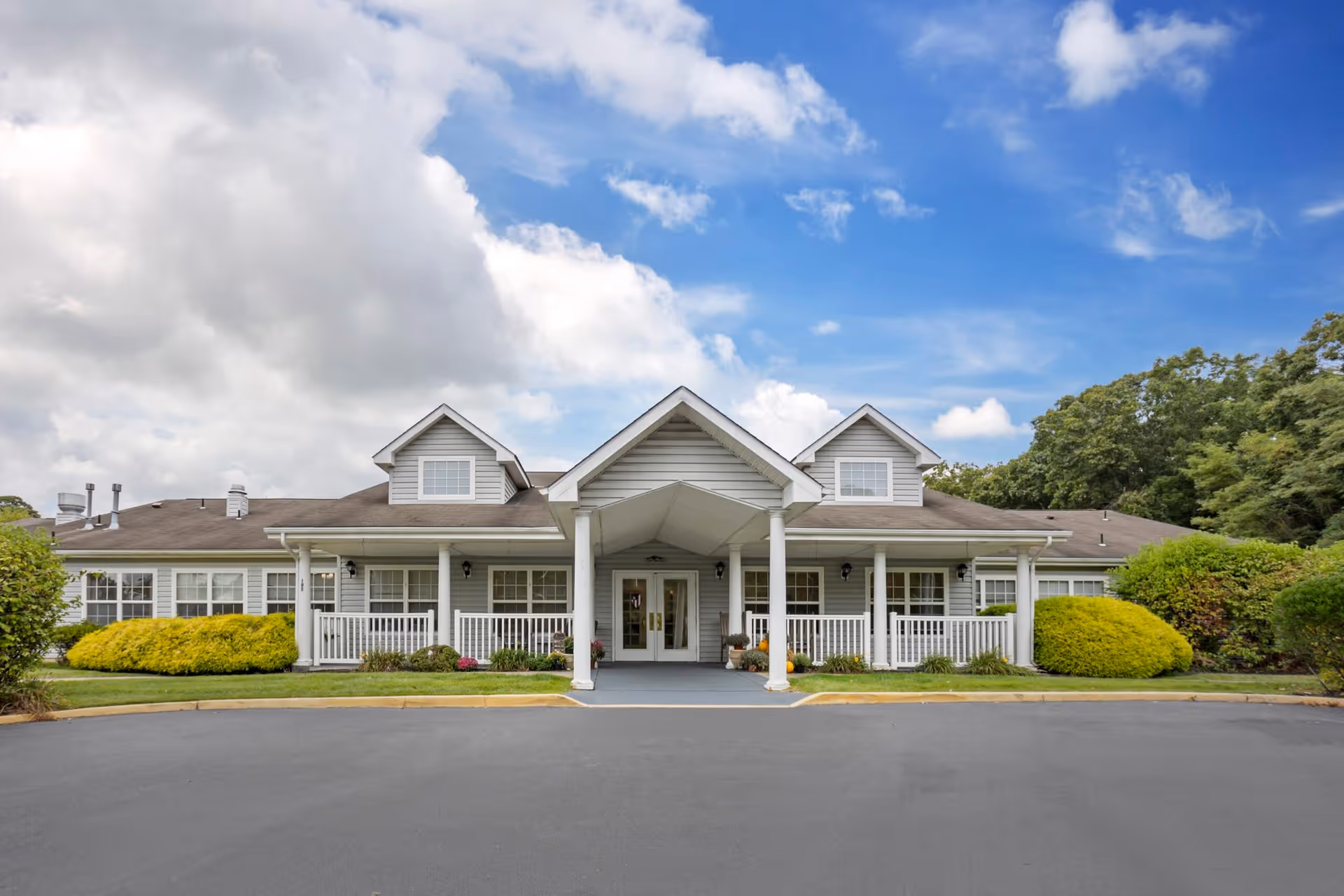 Front exterior view of a single-story building with a covered entrance supported by white columns, surrounded by green bushes and trees under a partly cloudy blue sky.