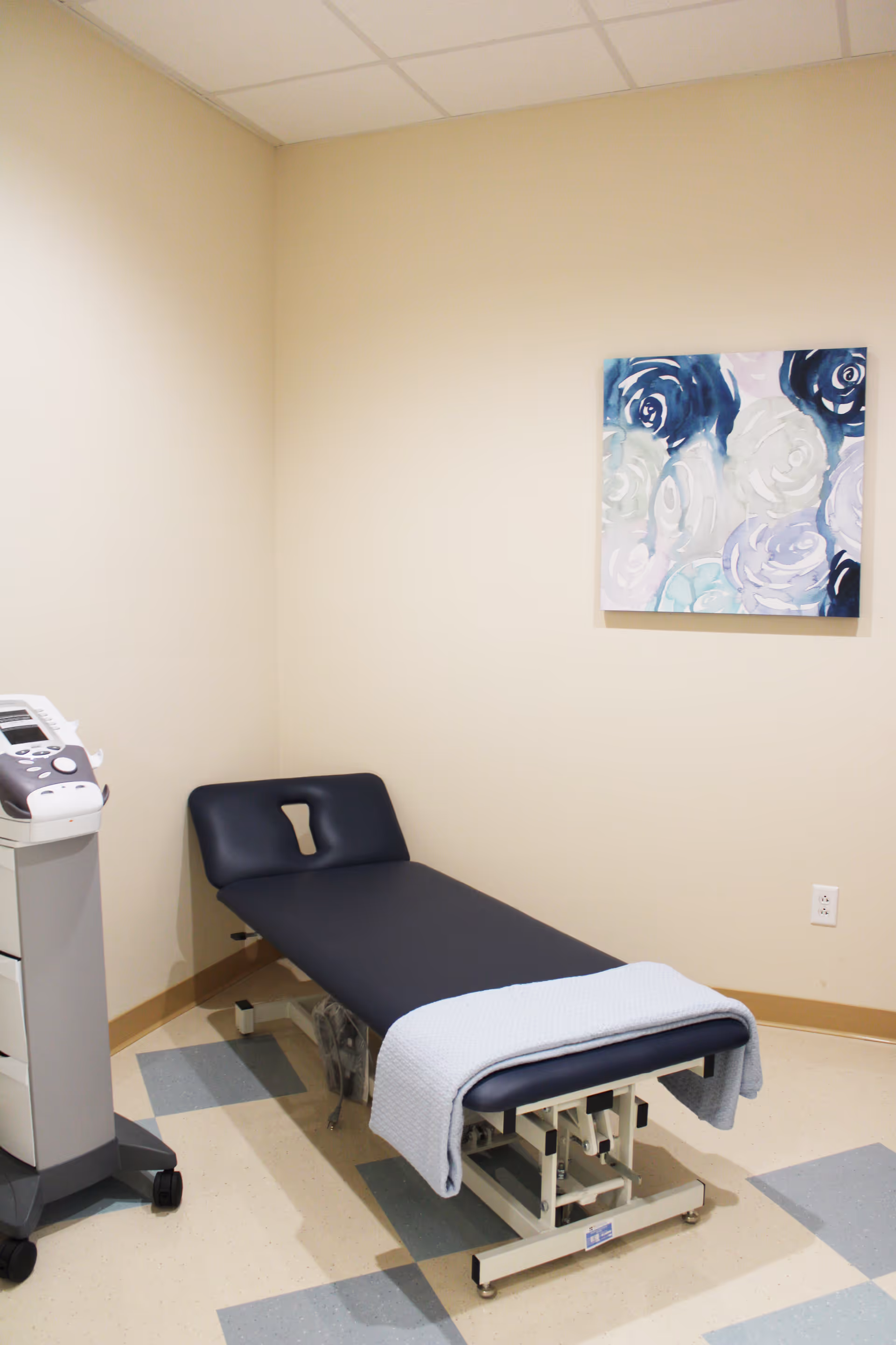 A small medical or therapy room with a black examination or therapy table covered partially with a light blue blanket. There is a medical device on a stand with wheels next to the table. The walls are beige, and a colorful abstract painting with blue and white tones hangs on the wall.