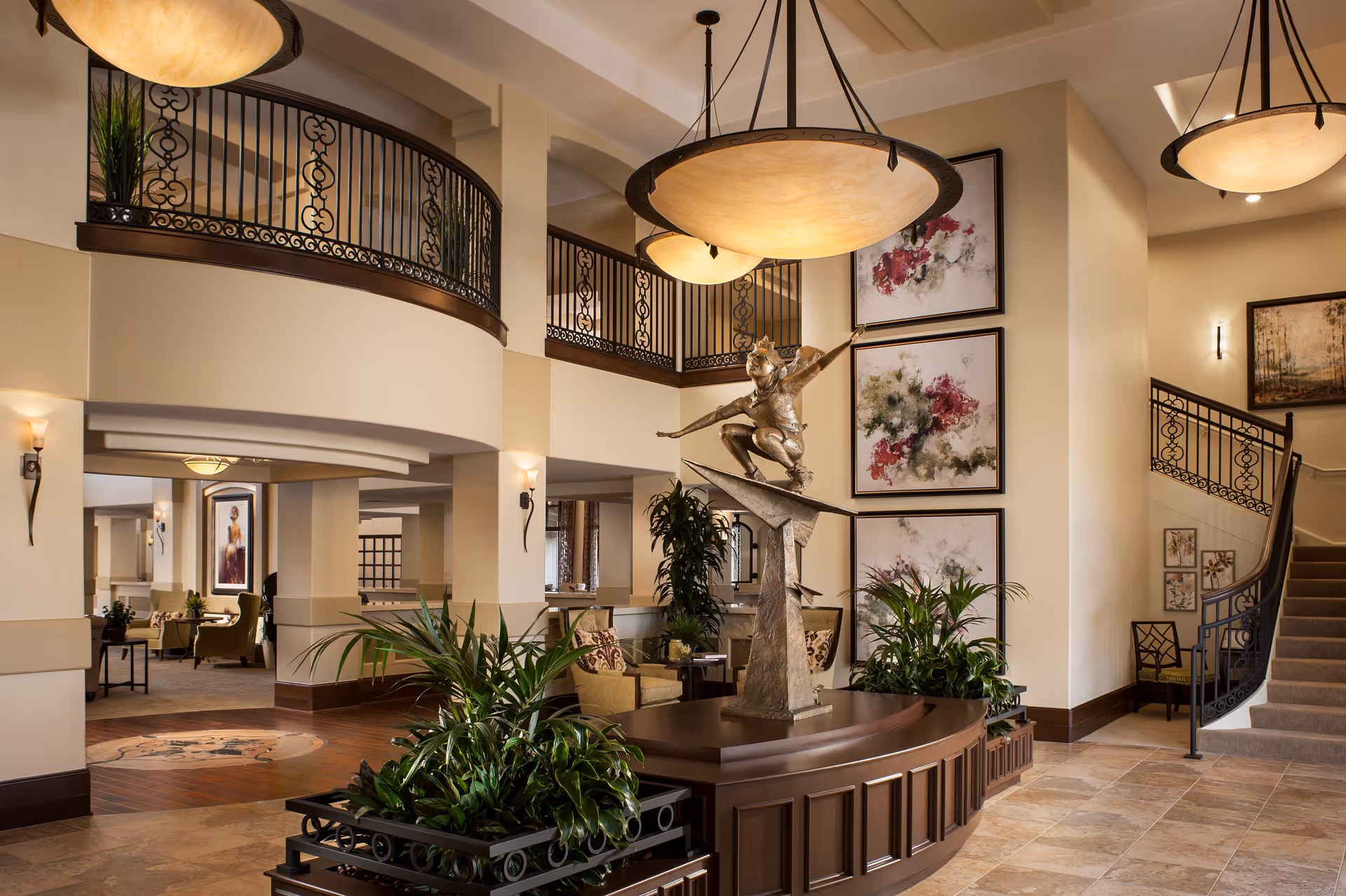 Spacious and elegant interior of a senior living facility featuring a bronze sculpture of a child on a pedestal surrounded by plants. The area has warm lighting with large hanging lamps, decorative wrought iron railings on the upper balcony and staircase, and abstract floral paintings on the walls. Comfortable seating areas are visible in the background.