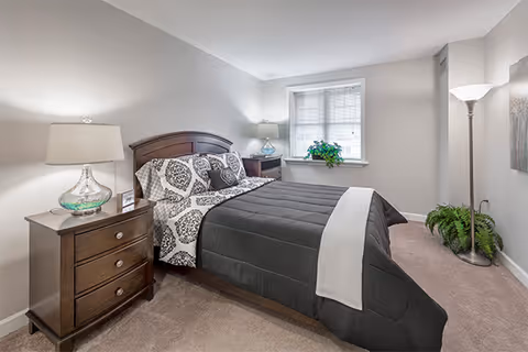 A neatly made bed with a dark gray comforter and patterned pillows in a bedroom. There is a wooden nightstand with a glass lamp on the left side of the bed and another small table with a lamp and a plant near the window. The room has beige carpet, light gray walls, a window with blinds, and a floor lamp with a white shade in the corner.