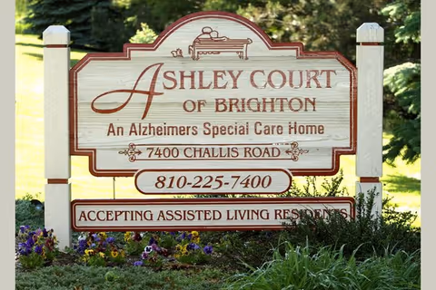Wooden sign for Ashley Court of Brighton, an Alzheimer's Special Care Home, located at 7400 Challis Road with phone number 810-225-7400, surrounded by grass and flowers.