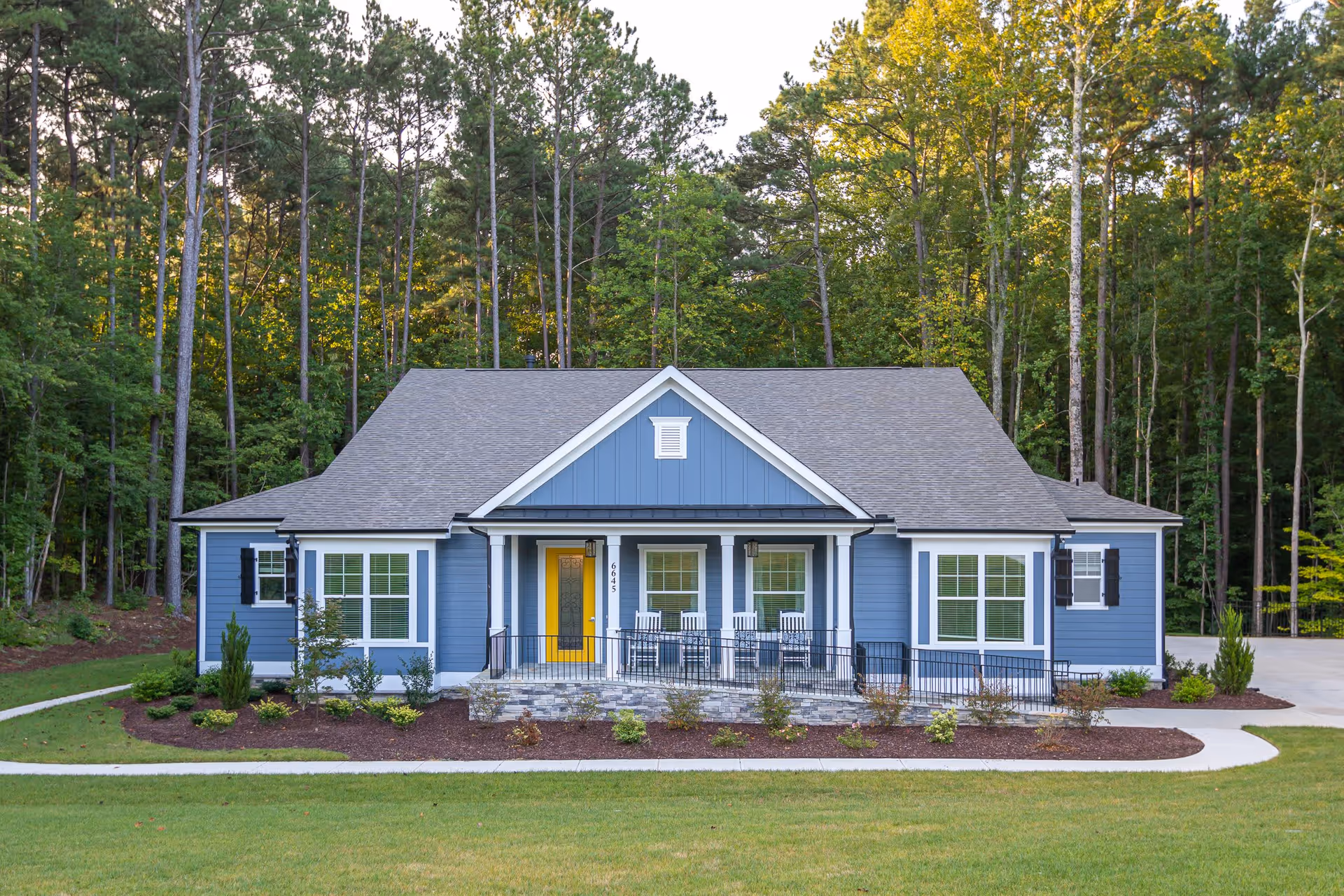 Front exterior view of a single-story blue house with a gray roof, a yellow front door, white-framed windows, and a porch with four white rocking chairs. The house is surrounded by a well-maintained lawn and landscaping, with tall trees in the background.