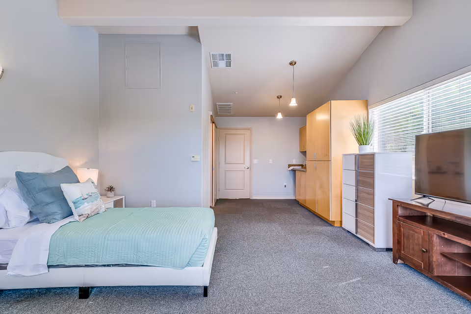 A spacious senior living bedroom with a white bed frame, light blue bedding, and decorative pillows. There is a nightstand with a lamp and a small plant next to the bed. Across from the bed is a wooden TV stand with a flat-screen TV and a tall dresser with a potted plant on top. The room has gray carpet, light-colored walls, and a large window with blinds letting in natural light. The entrance door and a kitchenette area with wooden cabinets are visible in the background.