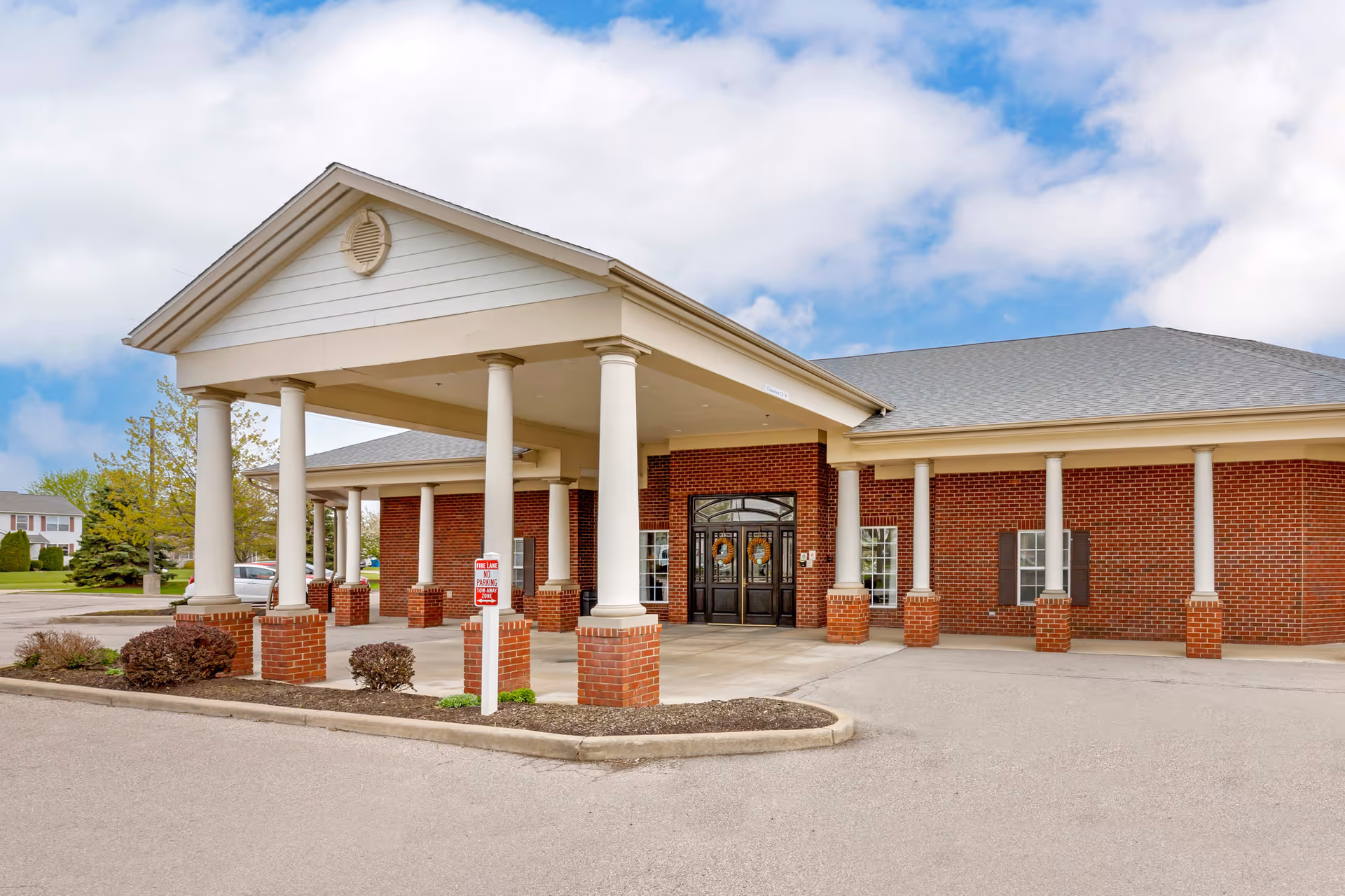 Front exterior view of a single-story brick building with white columns supporting a covered entrance. The building has a gray shingled roof and black double doors at the entrance. There are small landscaped areas with bushes near the entrance and a sign indicating no parking in the fire lane. The sky is partly cloudy.
