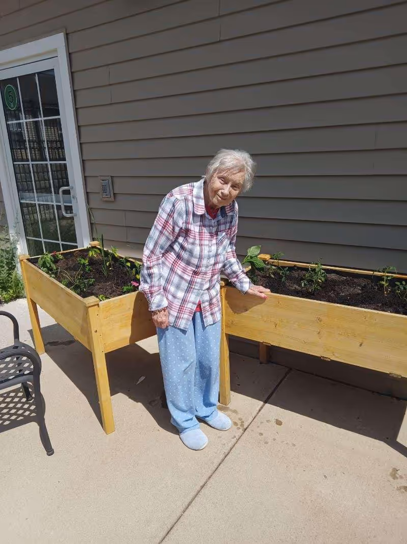 An elderly woman wearing a plaid shirt and blue pajama pants stands outside next to two wooden raised garden beds with small plants growing in them. The setting appears to be a patio area with a building wall and a glass door in the background.