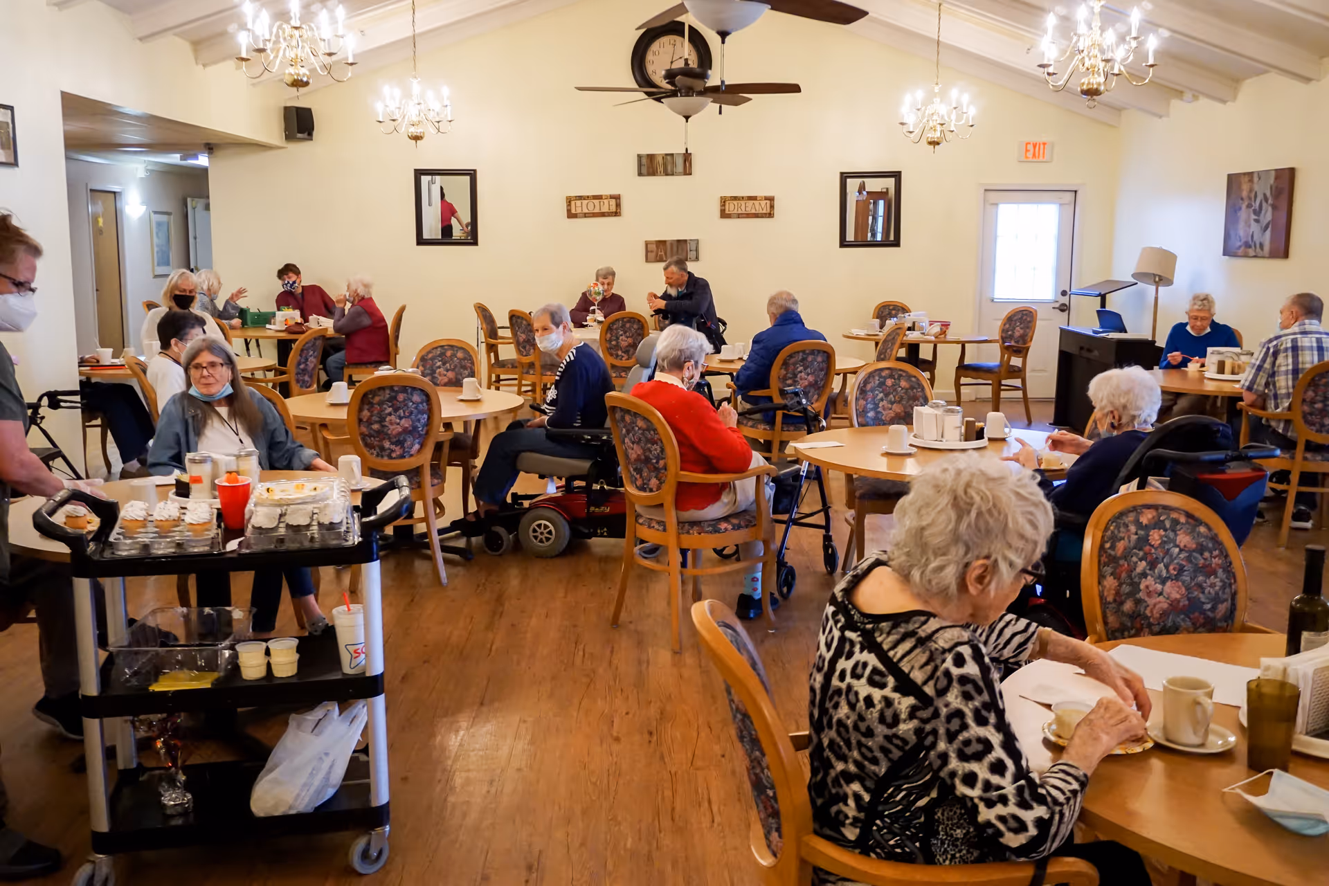 A dining room in an assisted living facility with elderly residents seated at round tables, some wearing masks. A staff member is pushing a cart with food and drinks. The room has chandeliers, ceiling fans, and wall decorations with words like 'Hope', 'Dream', and 'Faith'.