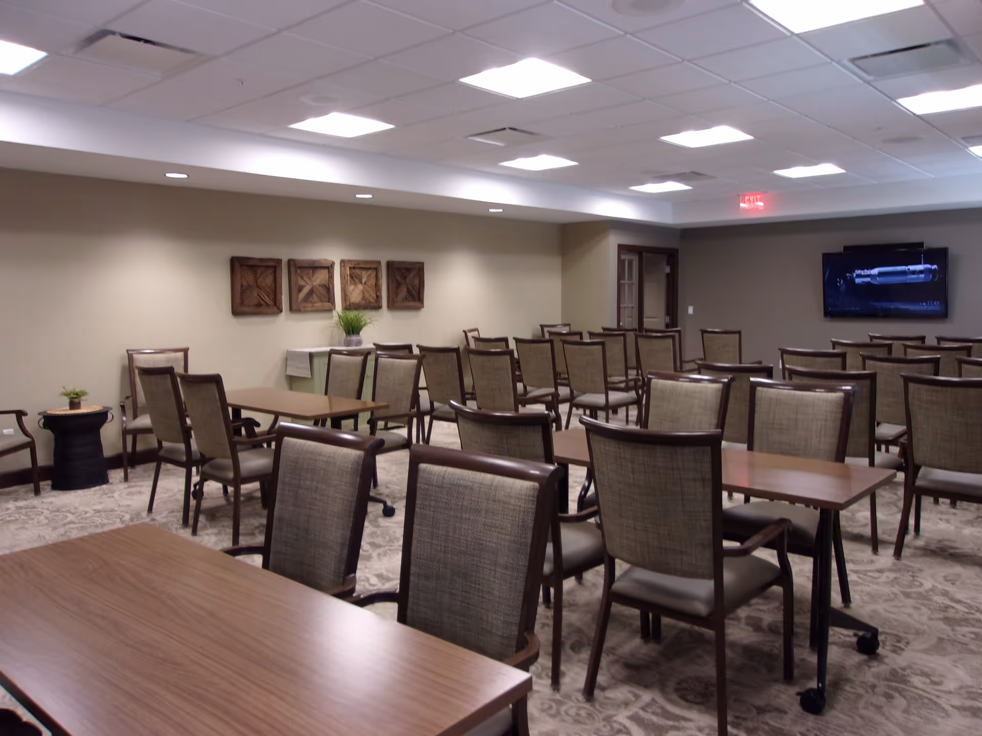 A well-lit room with multiple rows of wooden tables and cushioned chairs arranged in a classroom or meeting style. The walls are beige with four square wooden wall decorations and a small table with a plant. A flat-screen TV is mounted on the far wall, and there is an exit sign above a door in the back corner.